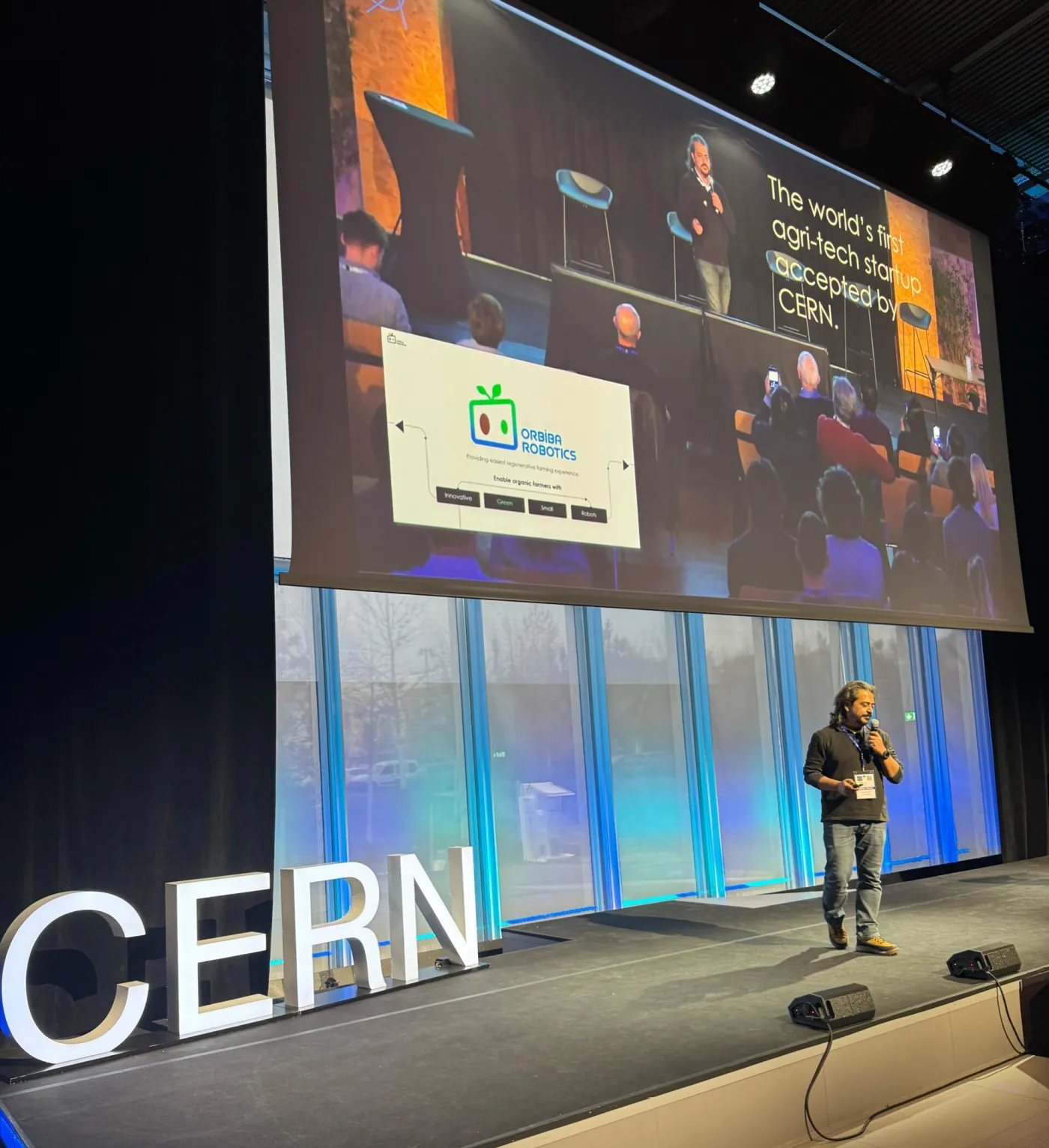 İlker Bektaş CERN sahnesinde, arkasındaki ekranda Orbiba Robotics sunumuyla konuşuyor